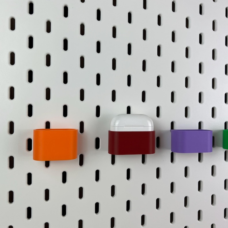 May include: A white pegboard with vertical slots features several small, colorful plastic storage containers attached to it. One container is bright orange, one is deep red holding white wireless earbuds, and one is purple, all mounted securely.