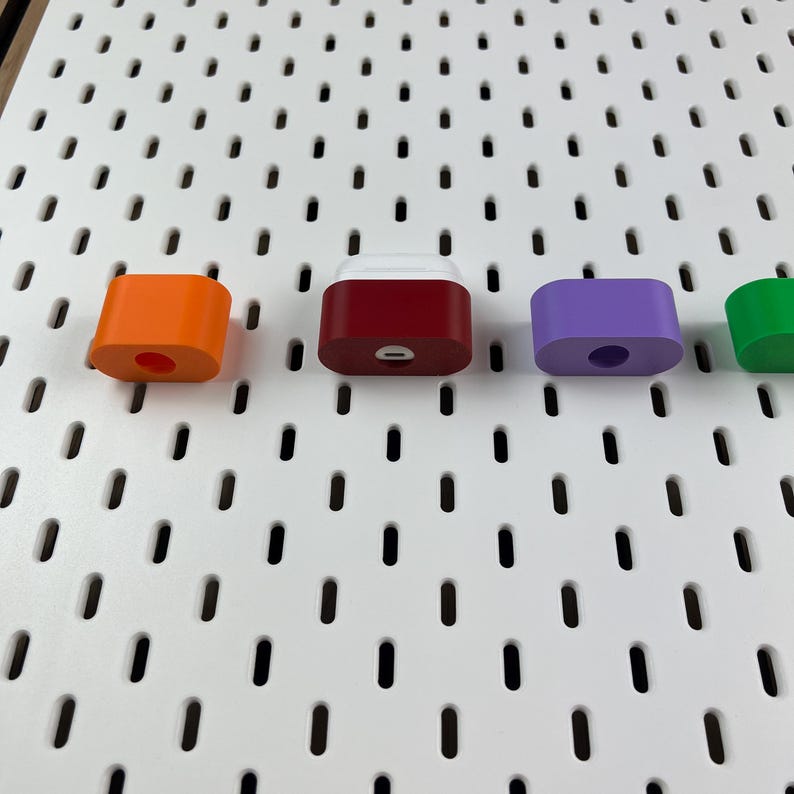 May include: A row of four colorful silicone charging cases for wireless earbuds, featuring an orange, a dark red, a purple, and a green case. Each case has a small circular or oval cutout on the front, and the dark red case shows a charging port.