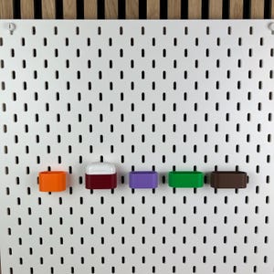 May include: A white pegboard with vertical slots features five small, rectangular storage containers attached to it. The containers are colored orange, dark red, purple, green, and brown. One dark red container holds a white wireless earbud case.