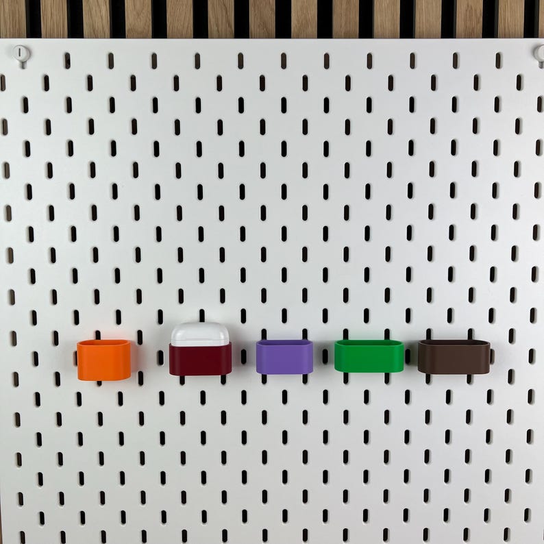 May include: A white pegboard with vertical slots features five small, rectangular storage containers attached to it. The containers are colored orange, dark red, purple, green, and brown. One dark red container holds a white wireless earbud case.