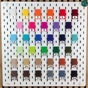 May include: A white rectangular pegboard featuring thirty-four small, rectangular, solid-colored attachments arranged in six rows. Each attachment is labeled with a white circular sticker numbered from 1 to 34, displaying a wide spectrum of colors.