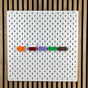 May include: A white rectangular pegboard with a grid of vertical slots features five small, colorful storage attachments mounted in a horizontal row. The attachments are orange, white and red, purple, green, and brown, providing modular organization.