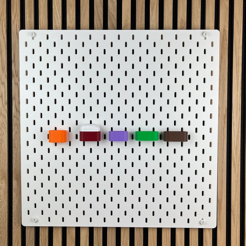 May include: A white rectangular pegboard with a grid of vertical slots features five small, colorful storage attachments mounted in a horizontal row. The attachments are orange, white and red, purple, green, and brown, providing modular organization.