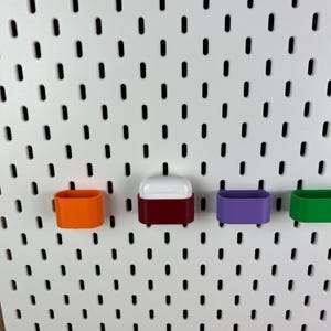 May include: A white pegboard wall organizer featuring four small, rectangular storage bins in orange, maroon, purple, and green. The maroon bin contains a white wireless earbud charging case, while the other three bins remain empty.