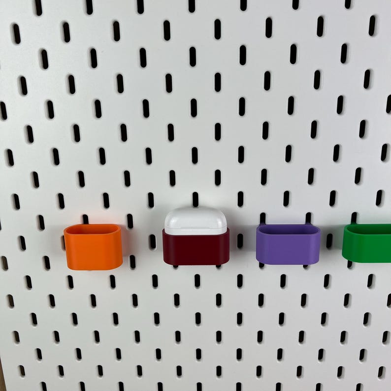 May include: A white pegboard wall organizer featuring four small, rectangular storage bins in orange, maroon, purple, and green. The maroon bin contains a white wireless earbud charging case, while the other three bins remain empty.