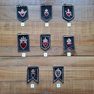 Op de afbeelding: Acht zwarte stoffen wimpelspelden gelabeld van A tot H, elk voorzien van een zilverkleurig metalen heilig hart-embleem met rode accenten. Elke wimpel is bevestigd aan een zilveren veiligheidsspeld en versierd met witte stiksels en details.