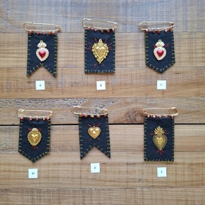 May include: Six black fabric pennant pins labeled A through F, each featuring a gold-tone metal heart charm, red embroidered rays, and small red beads along the top safety pin. The fabric edges are stitched with yellow thread.