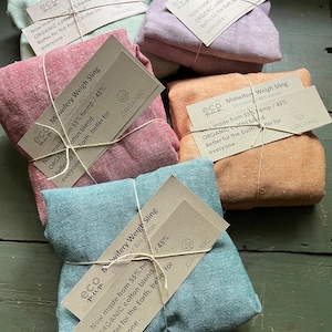 May include: Five folded fabric weigh slings in light green, lavender, red, orange, and blue. Each sling is wrapped in twine with a tan paper tag labelled Eco Mum Midwifery Weigh Sling, stating it is a 55% hemp and 45% organic cotton blend.