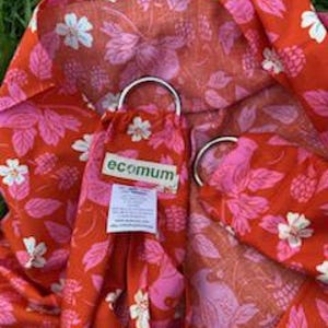 Könnte beinhalten: Eine rot-rosa Babytrage mit Blumenmuster und zwei silbernen Ringen. Die Trage hat ein Muster aus Blumen und Beeren. Der Markenname "ecomum" ist auf einem am Träger befestigten Etikett zu sehen.