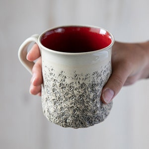 May include: A white ceramic mug with a red interior and a textured, speckled exterior. The mug is being held by a hand.
