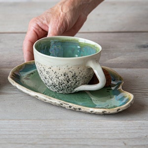 May include: Two ceramic teacups with speckled glaze and matching saucers. The cups are teal and green with white interiors. One saucer has a chocolate truffle on it.