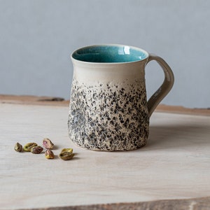 Pode incluir: Uma caneca de cerâmica branca salpicada com um interior azul turquesa e uma alça. A caneca está sobre uma superfície de madeira clara com pistache espalhados ao redor.