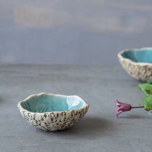 May include: Three small ceramic bowls with a light blue glaze and a textured white exterior. The bowls are arranged on a gray surface with a sprig of purple flowers.