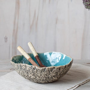 May include: A turquoise ceramic bowl with a speckled brown glaze and a rough, textured exterior. Two wooden spoons are resting inside the bowl. The bowl is sitting on a linen napkin on a wooden table.