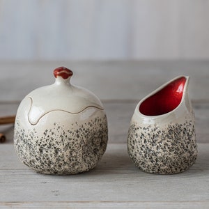 Könnte beinhalten: Eine weiße Zuckerdose aus Keramik mit rotem Deckel und einem passenden Milchkännchen. Beide Teile haben eine gesprenkelte schwarz-weiße Glasur.