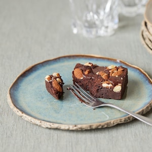 Könnte beinhalten: Ein Stück Schokoladen-Brownie mit Mandeln auf einem hellblauen Teller mit rustikalem Rand. Ein kleines Brownie-Stück und eine Gabel befinden sich ebenfalls auf dem Teller. Weitere Teller sind im Hintergrund gestapelt.