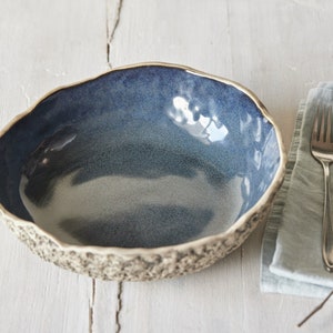 Könnte beinhalten: Eine blaue Keramikschüssel mit einem strukturierten Rand und einem glatten, glänzenden Innenraum. Die Schüssel steht auf einer weißen Holzoberfläche neben einer blauen Leinenserviette, einer Gabel und einem Löffel.