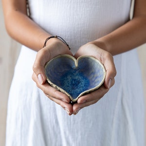May include: A heart-shaped ceramic bowl with a blue and white glaze. The bowl is being held in the hands of a person.