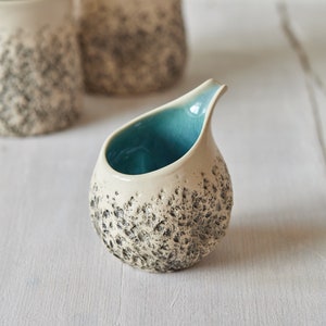 Puede incluir: Un peque&ntilde;o creamer de cer&aacute;mica blanca con textura moteada y un interior azul turquesa. El creamer tiene una forma redondeada y un peque&ntilde;o pico.