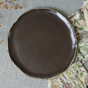 May include: A dark brown ceramic plate with a slightly irregular rim. The plate is sitting on a light gray textured surface with a floral patterned fabric napkin to the right.