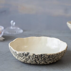 May include: A white ceramic bowl with a speckled black and gray glaze. The bowl has a wide, shallow shape and a slightly rippled edge.