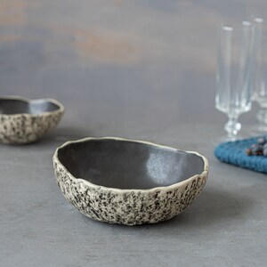 May include: Two ceramic bowls with a speckled, textured exterior and a dark gray interior. The bowls are on a gray surface.