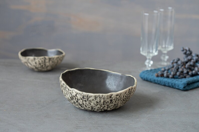 May include: Two ceramic bowls with a speckled, textured exterior and a dark gray interior. The bowls are on a gray surface.