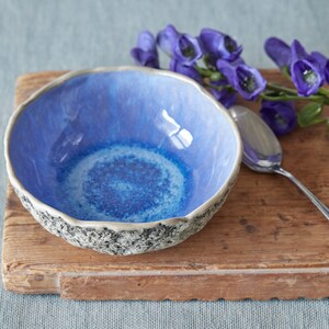 May include: A blue ceramic bowl with a textured rim sits on a wooden surface. A silver spoon and purple flowers are also on the surface.