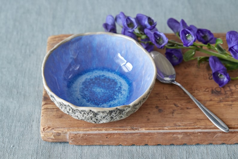 May include: A blue ceramic bowl with a textured rim sits on a wooden surface. A silver spoon and purple flowers are also on the surface.