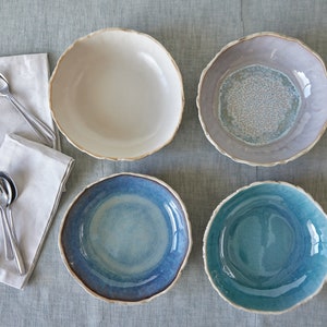 May include: Four ceramic bowls in different colours and patterns. The bowls are white, light blue, dark blue, and teal. They are all round and have a slightly uneven rim.