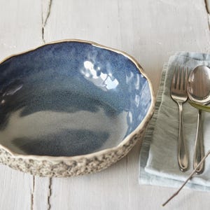 May include: A blue ceramic bowl with a textured rim and a light blue glaze. The bowl is sitting on a white wooden surface. A blue linen napkin with a silver fork and spoon is to the right of the bowl.