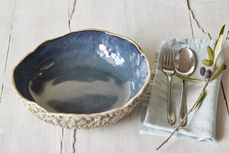 May include: A blue ceramic bowl with a textured rim and a light blue glaze. The bowl is sitting on a white wooden surface. A blue linen napkin with a silver fork and spoon is to the right of the bowl.