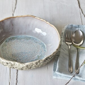 May include: A light blue and gray ceramic bowl with a textured rim and a speckled interior. The bowl is sitting on a white wooden surface next to a blue linen napkin with silver silverware and an olive branch.