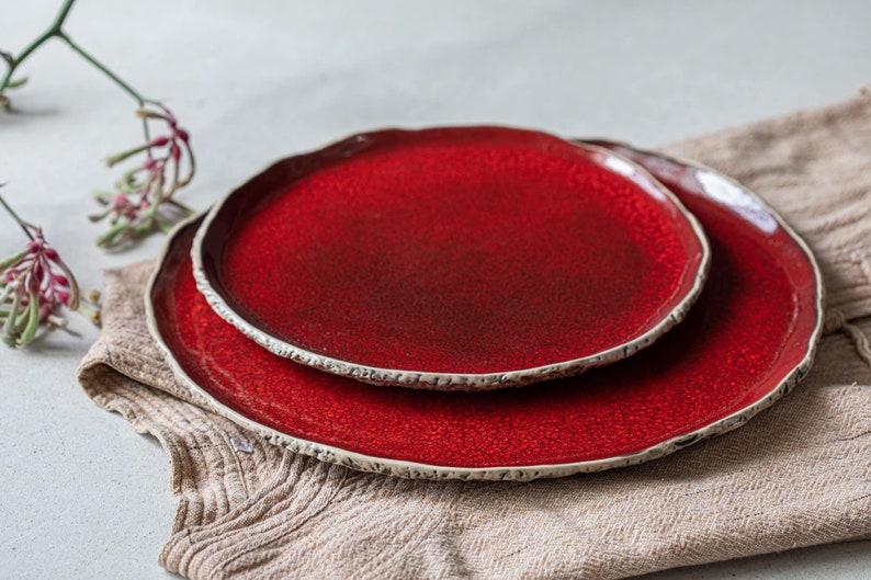 May include: Two red ceramic plates with a textured rim. The plates are stacked on top of each other and are sitting on a beige fabric surface.