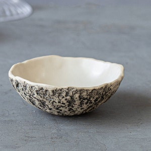 May include: A small, white ceramic bowl with a textured, black and white speckled exterior. The bowl is round and has a slightly flared rim.