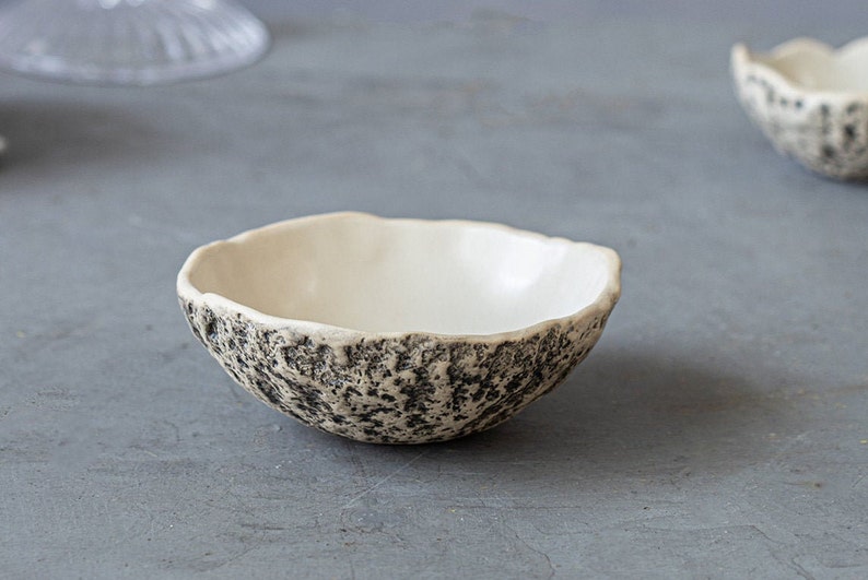 May include: A small, white ceramic bowl with a textured, black and white speckled exterior. The bowl is round and has a slightly flared rim.