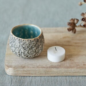 May include: A small, teal and gray ceramic candle holder with a textured surface sits on a wooden cutting board with a white tea light candle.