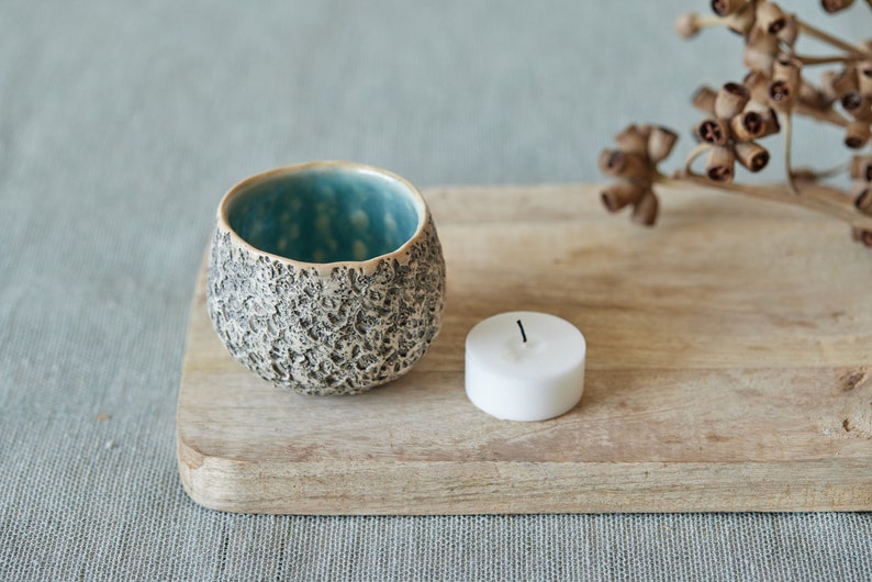 May include: A small, teal and gray ceramic candle holder with a textured surface sits on a wooden cutting board with a white tea light candle.