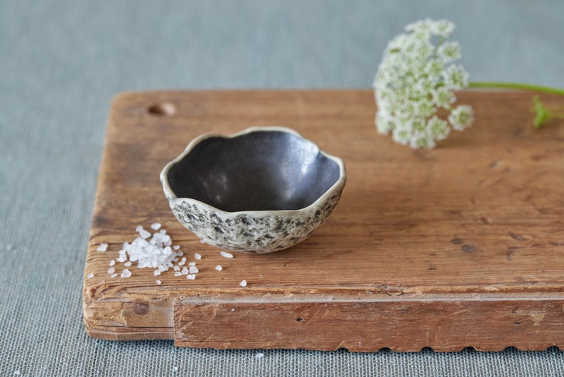 May include: A small, black and white speckled ceramic bowl sits on a wooden chopping board. The bowl has a scalloped edge and is filled with white salt crystals.