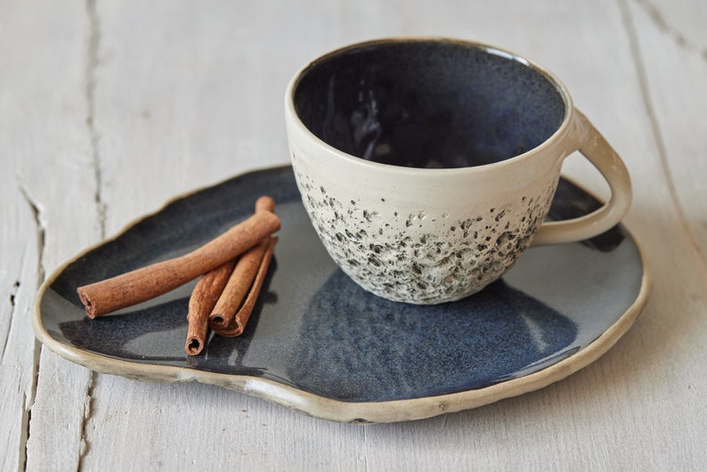 May include: A blue and white ceramic teacup and saucer set. The cup has a textured exterior and the saucer has a slightly irregular shape. Three cinnamon sticks are on the saucer.