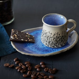 Puede incluir: Una taza de café de cerámica azul y blanca con un exterior texturizado se asienta sobre un platillo a juego. Un trozo de chocolate negro con chispas blancas y verdes descansa sobre el platillo. Los granos de café están esparcidos sobre una superficie de madera oscura.