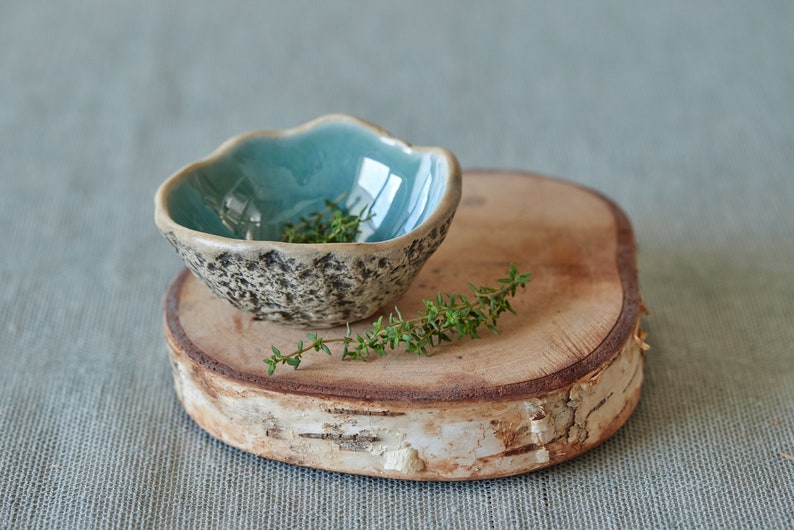May include: A small, teal blue ceramic bowl with a rough, textured exterior sits on a wooden slice. The bowl is filled with fresh green thyme sprigs.