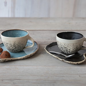 Pode incluir: Duas chávenas de chá de cerâmica com esmalte branco salpicado e interiores azuis e pretos contrastantes, sentadas em pratos de acompanhamento combinando. Um prato tem duas trufas de chocolate em cima.