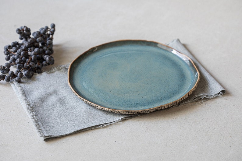 May include: A round ceramic plate with a glossy teal blue glaze and a textured, dark brown exposed clay rim. The plate rests on a rectangular grey linen napkin with frayed edges, accompanied by a small cluster of dark purple dried berries.