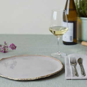 May include: A light gray ceramic dinner plate with a slightly uneven, textured rim. The plate is set on a light green tablecloth with a silver fork and knife on a gray linen napkin.