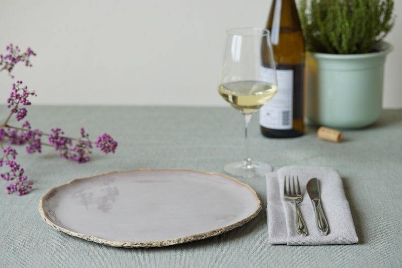 May include: A light gray ceramic dinner plate with a slightly uneven, textured rim. The plate is set on a light green tablecloth with a silver fork and knife on a gray linen napkin.