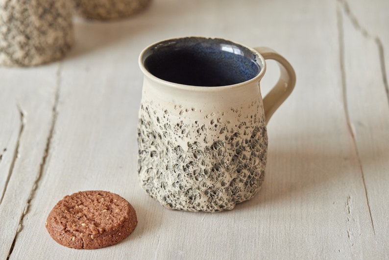 May include: A white ceramic mug with a textured, speckled surface and a dark blue interior. The mug has a handle and is sitting on a white wooden surface next to a chocolate chip cookie.