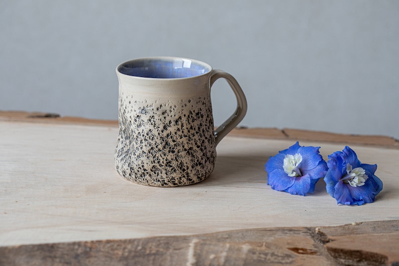 May include: A white ceramic mug with a speckled black glaze and a light blue interior. The mug has a rounded body and a handle. The mug is sitting on a wooden surface with two blue flowers.