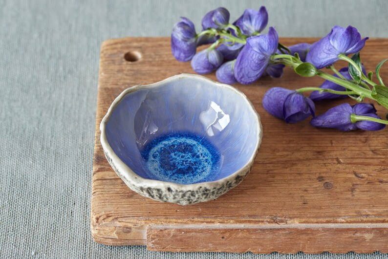 K&ouml;nnte beinhalten: Eine kleine, blaue und wei&szlig;e Keramiksch&uuml;ssel mit einem strukturierten Rand steht auf einem Holzbrett. Ein Zweig mit violetten Bl&uuml;ten liegt neben der Sch&uuml;ssel.