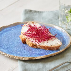 May include: A slice of toasted bread topped with white cream cheese and a mound of vibrant red beet sprouts, served on a round, textured blue ceramic plate with a rustic, uneven edge, accompanied by a silver fork and a glass of water.